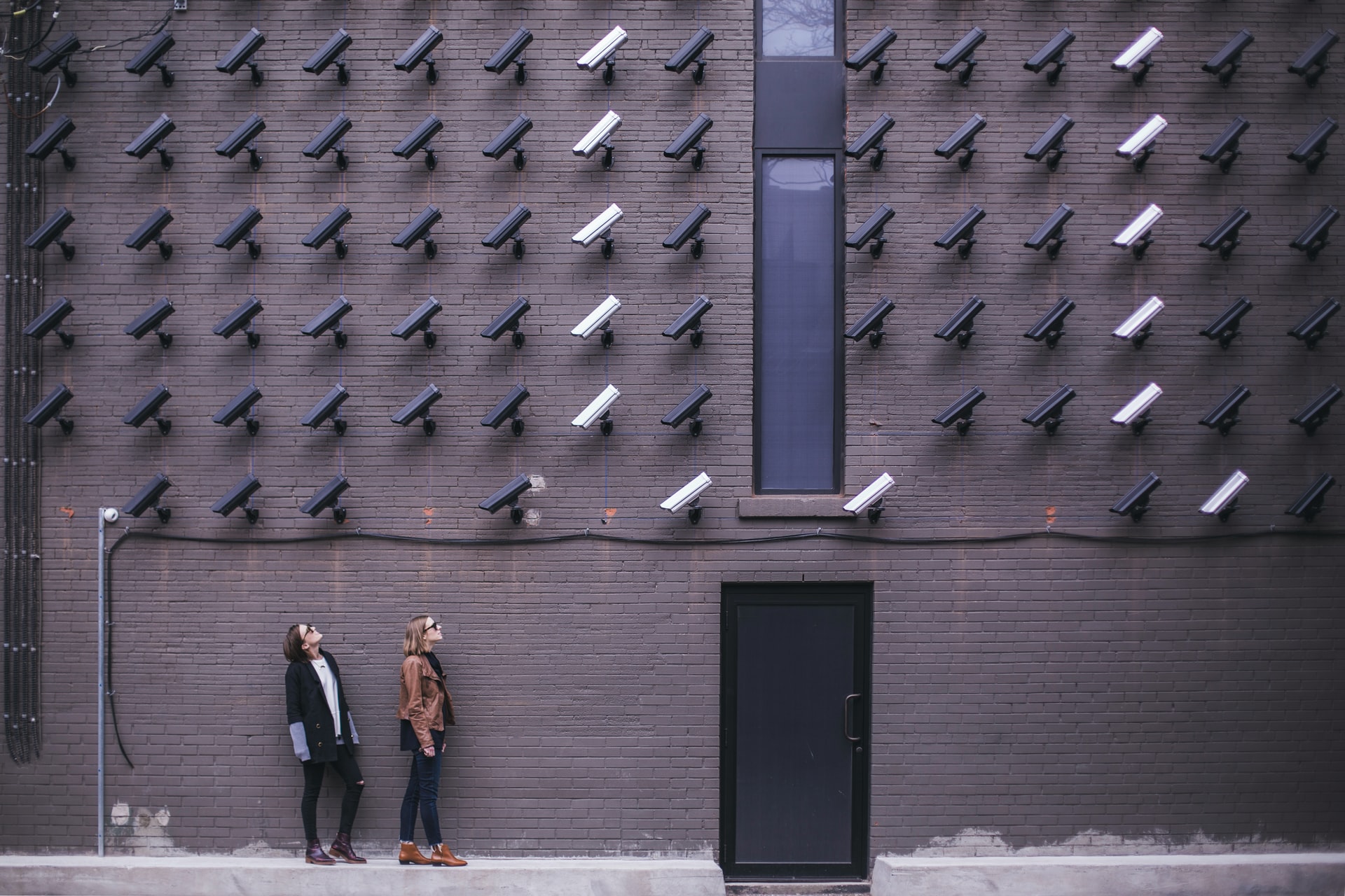 A large number of surveillence cameras on a wall. All are pointing in the same direction and are looking at two people.